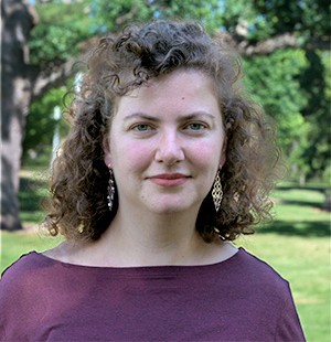 Head-and-shoulders portrait of Jeta Rudi Polloshka outdoors. The background shows green grass and trees with sunlight, softly blurred to keep focus on the subject
